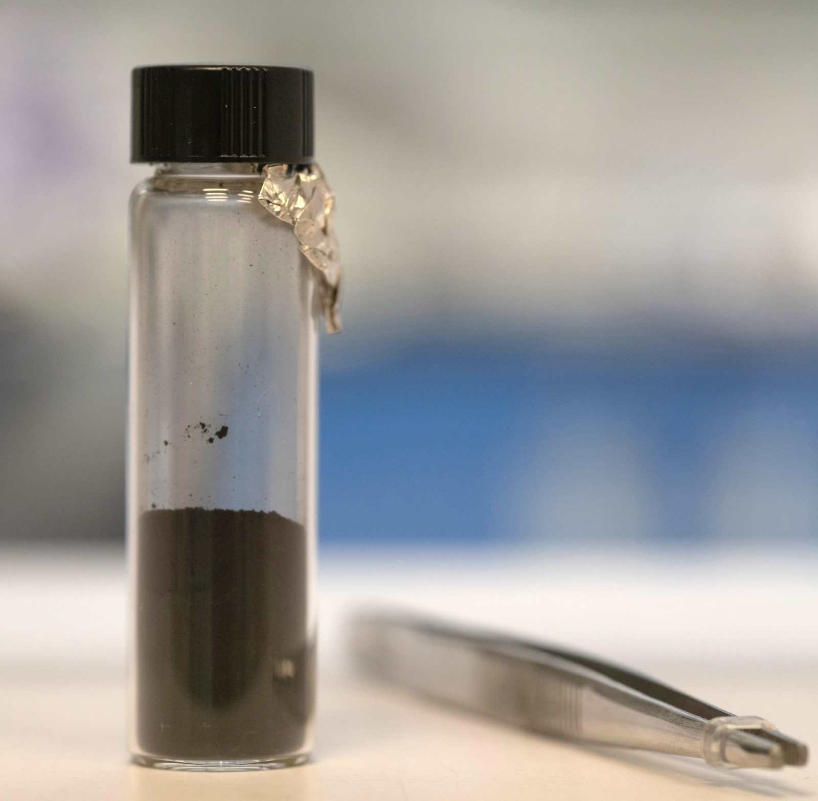 Un vial de vidrio con tapón negro, apoyado sobre una superficie clara, con un polvo oscuro y granuloso dentro.