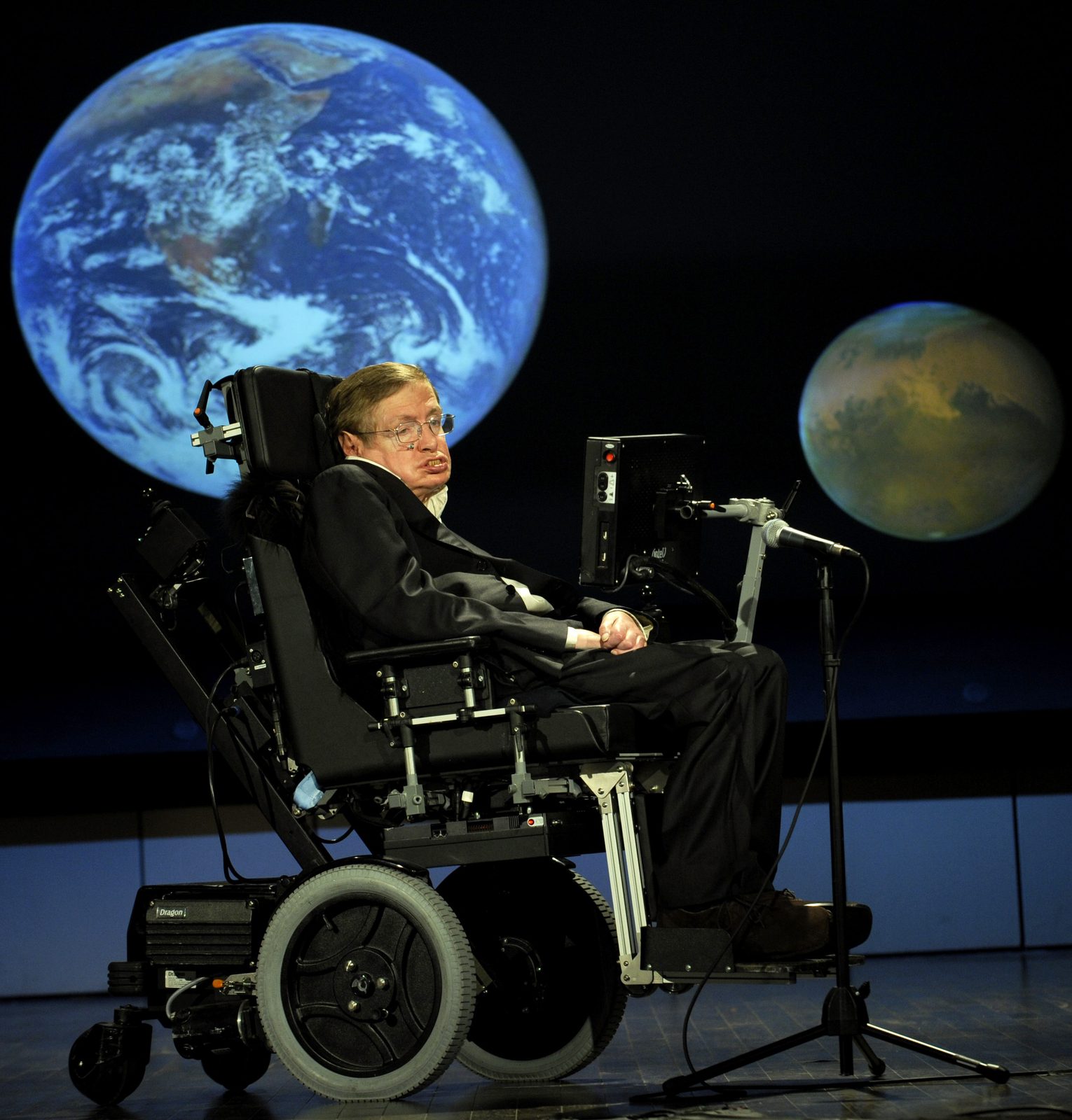 Stephen Hawking en su silla de ruedas ante un micrófono, en un escenario, con imágenes de la Tierra y de Marte proyectadas detrás.