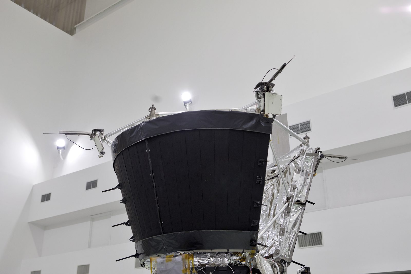 El cuerpo cónico de la sonda coronado por un escudo térmico circular oscuro, con antenas desplegadas, en una sala de procesamiento blanca.