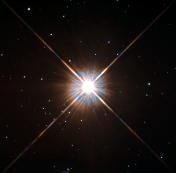 Un campo de estrellas sobre fondo negro con una estrella rojiza y brillante, de cruz de difracción marcada, cerca del centro.