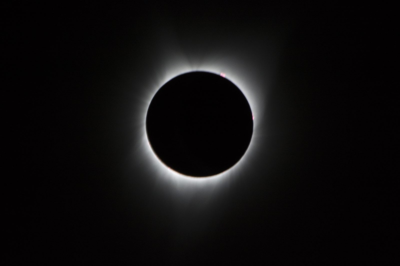 Un disco negro perfecto rodeado por una melena de luz blanca desflecada, con varios puntos rojos en el borde inferior.