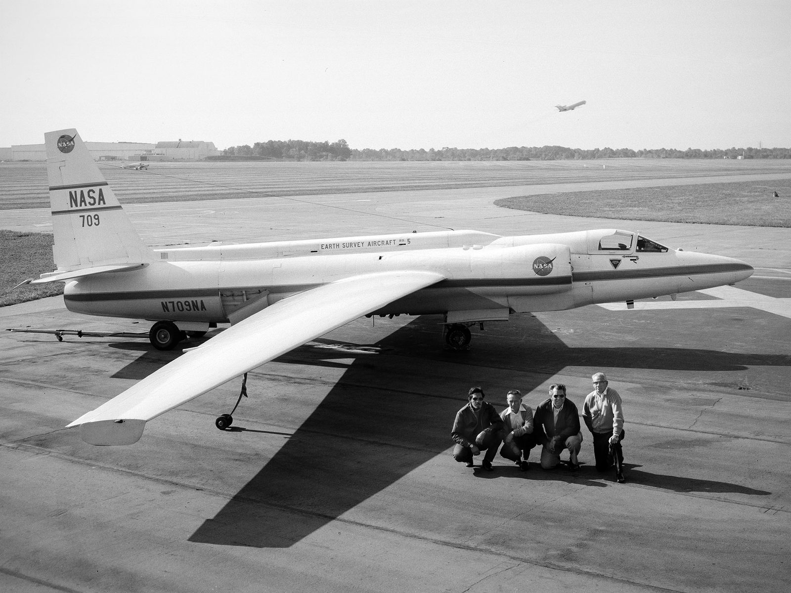Un avión blanco de alas muy largas y estrechas, estacionado en la pista, con cuatro hombres agachados delante y un hangar al fondo.