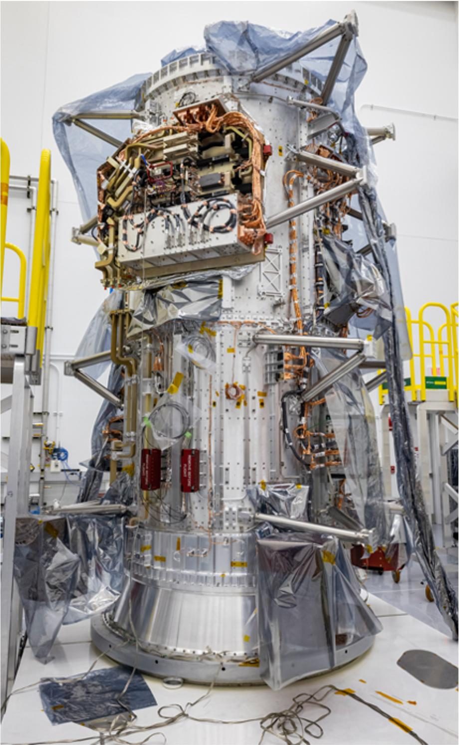 El cuerpo cilíndrico de la nave Europa Clipper, envuelto en plástico protector y erizado de tubos y cableado, de pie en una sala de montaje vacía.