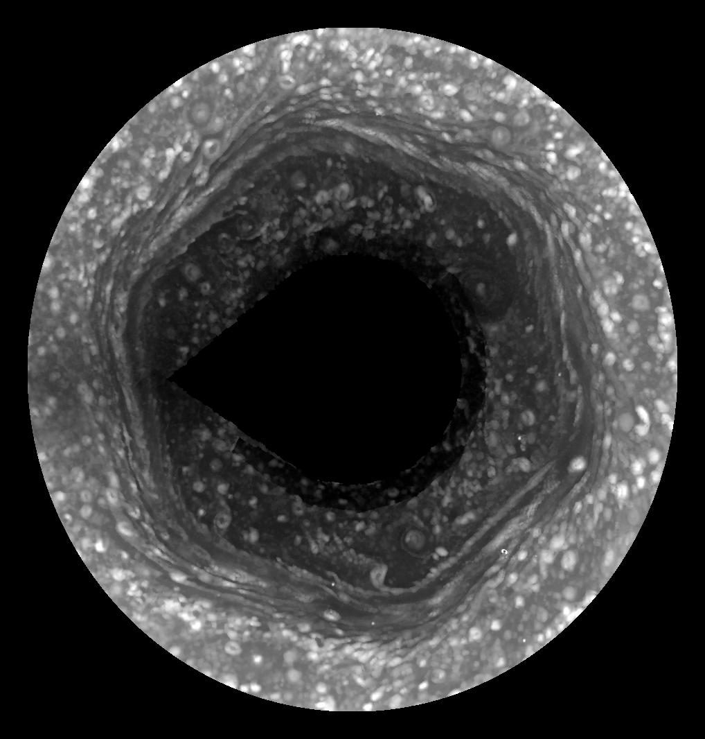 Vista cenital en blanco y negro del polo norte de Saturno, con bandas de nubes que dibujan un hexágono y un hueco negro sin datos en el centro.