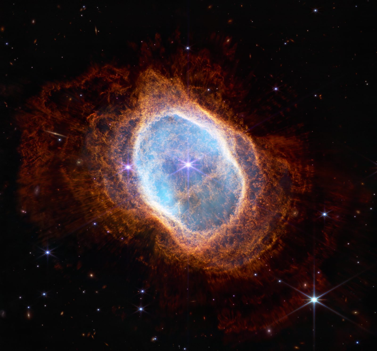 Una nebulosa ovalada de gas azul y naranja con dos estrellas en el centro, sobre un fondo sembrado de galaxias.