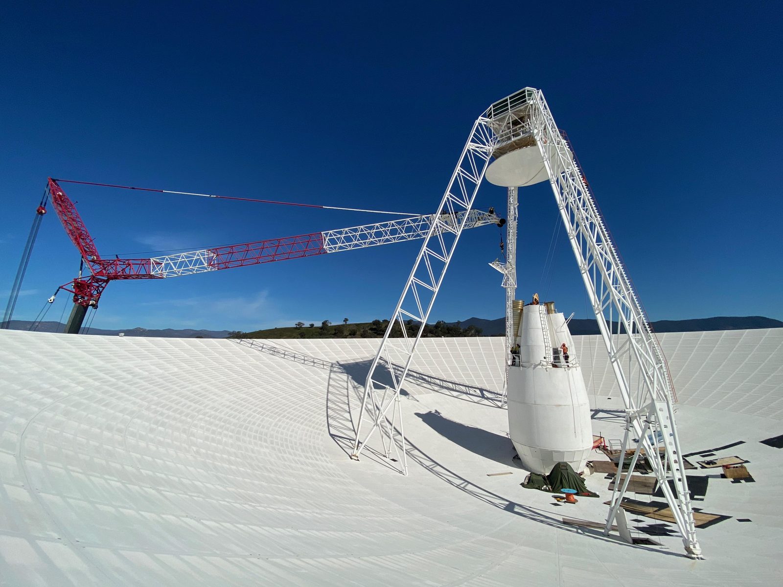 El interior de un plato de antena gigantesco, con un cono en el centro, una grúa roja y operarios diminutos trabajando en la estructura.