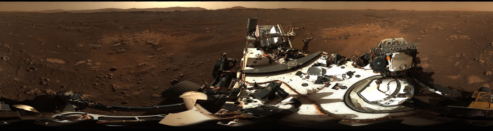 Panorama del suelo del cráter Jezero con la cubierta del rover Perseverance en primer plano: llanura de arena y piedras sueltas hasta un horizonte bajo, con el borde del cráter recortado a lo lejos.