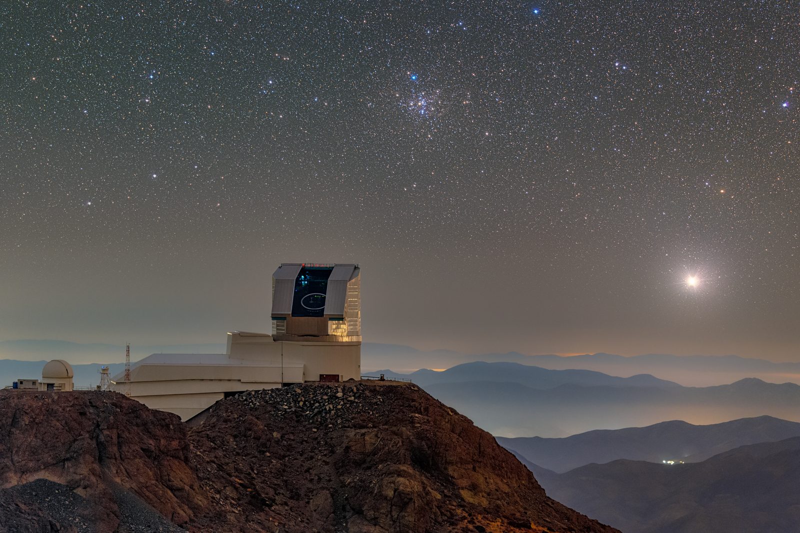 Edificio del observatorio en lo alto de un cerro, de noche, con la cúpula abierta y un cielo cuajado de estrellas encima.