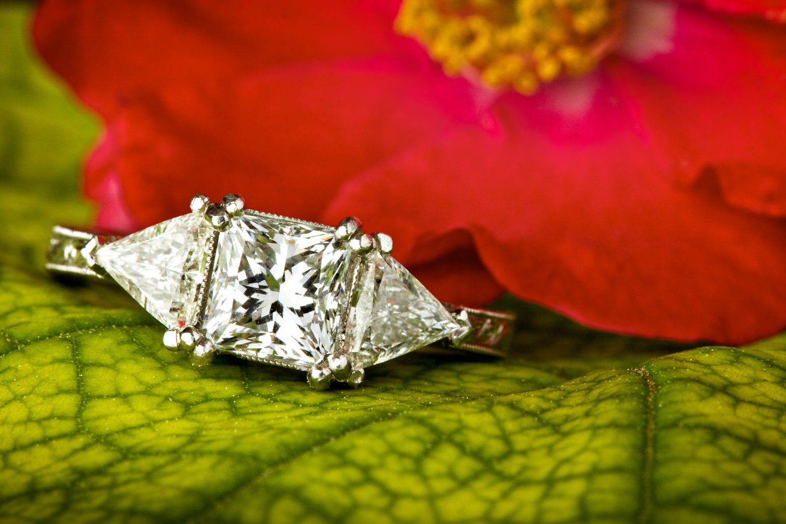 Macro de un anillo con un diamante central de talla cuadrada y dos triangulares a los lados, apoyado sobre una hoja verde, con una flor roja detrás.
