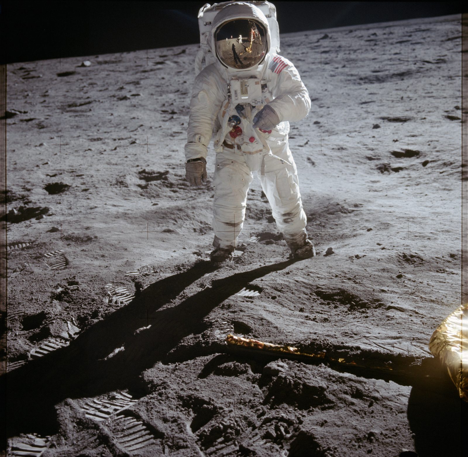 Un astronauta de pie sobre el suelo lunar, iluminado de lleno por el Sol, con el visor dorado bajado y el cielo completamente negro detrás.