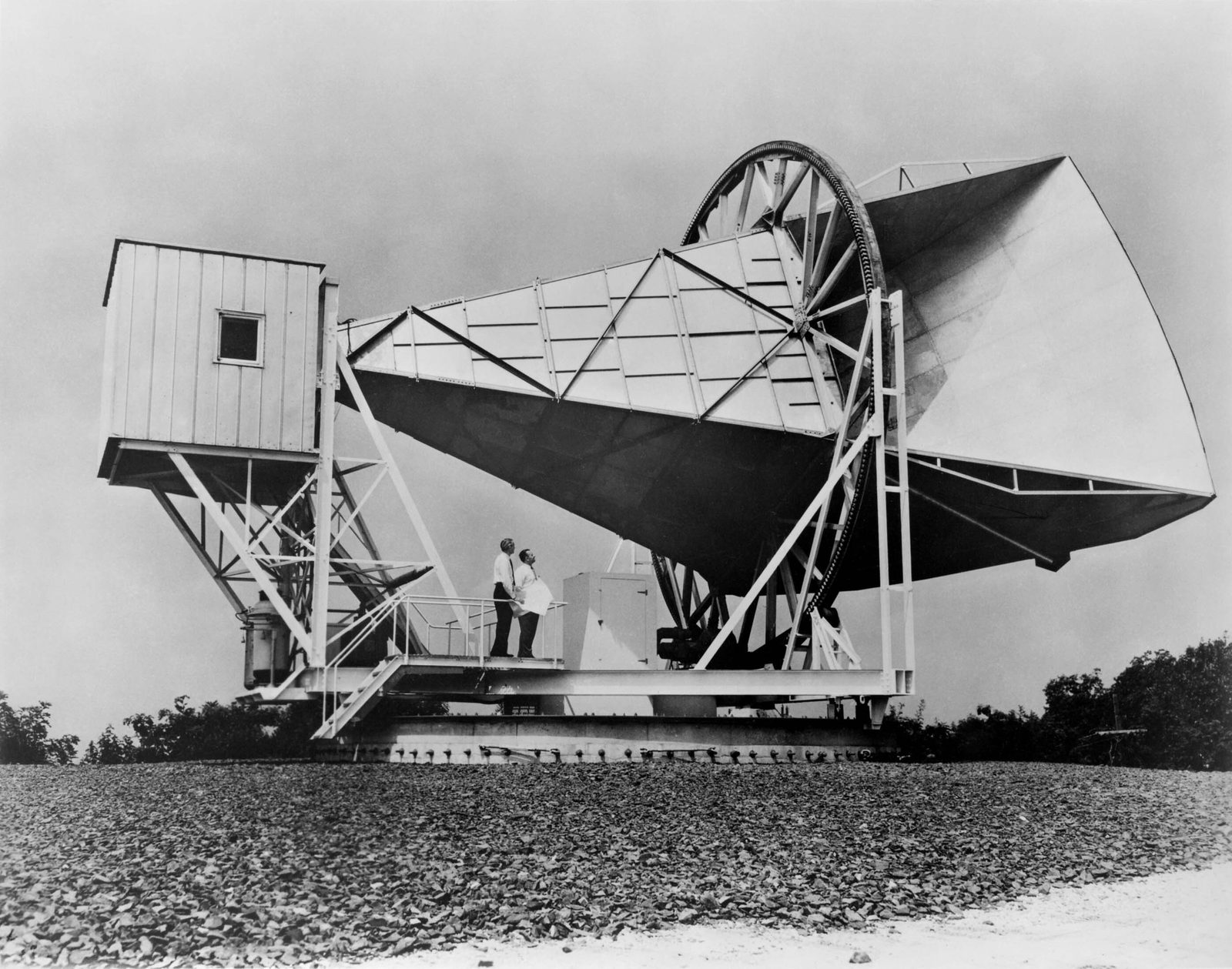 La antena de bocina de Holmdel, sobre grava plana, apuntando al cielo, con dos personas de pie en su plataforma.