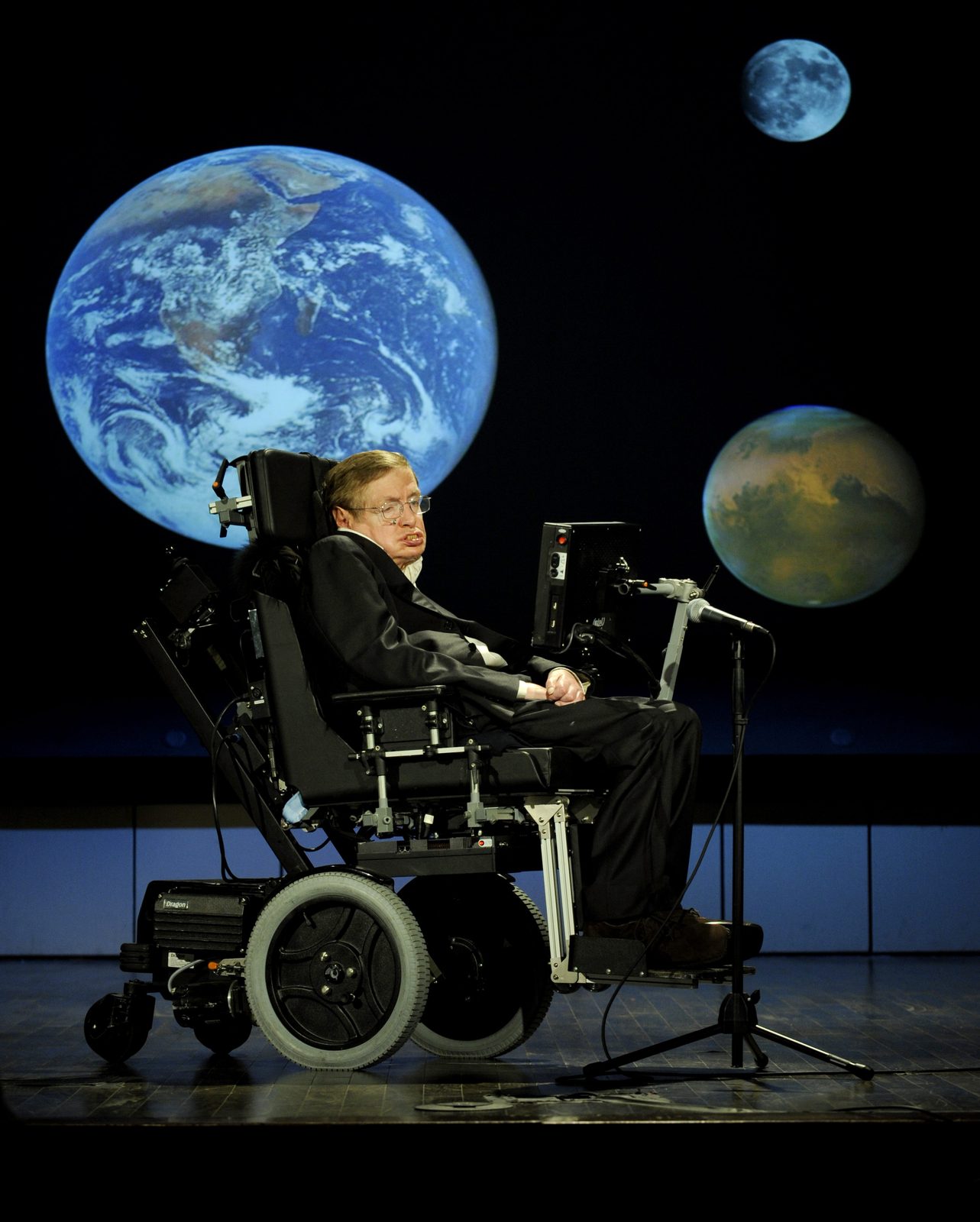Stephen Hawking en su silla, en primer plano, con una pantalla de proyección detrás que muestra la Tierra, la Luna y Marte.