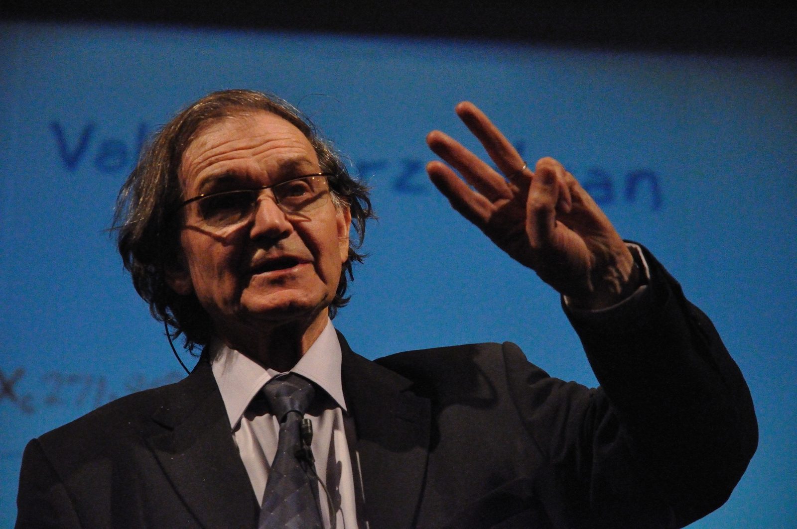 Roger Penrose de perfil tres cuartos, hablando, con una mano levantada que muestra dos dedos.