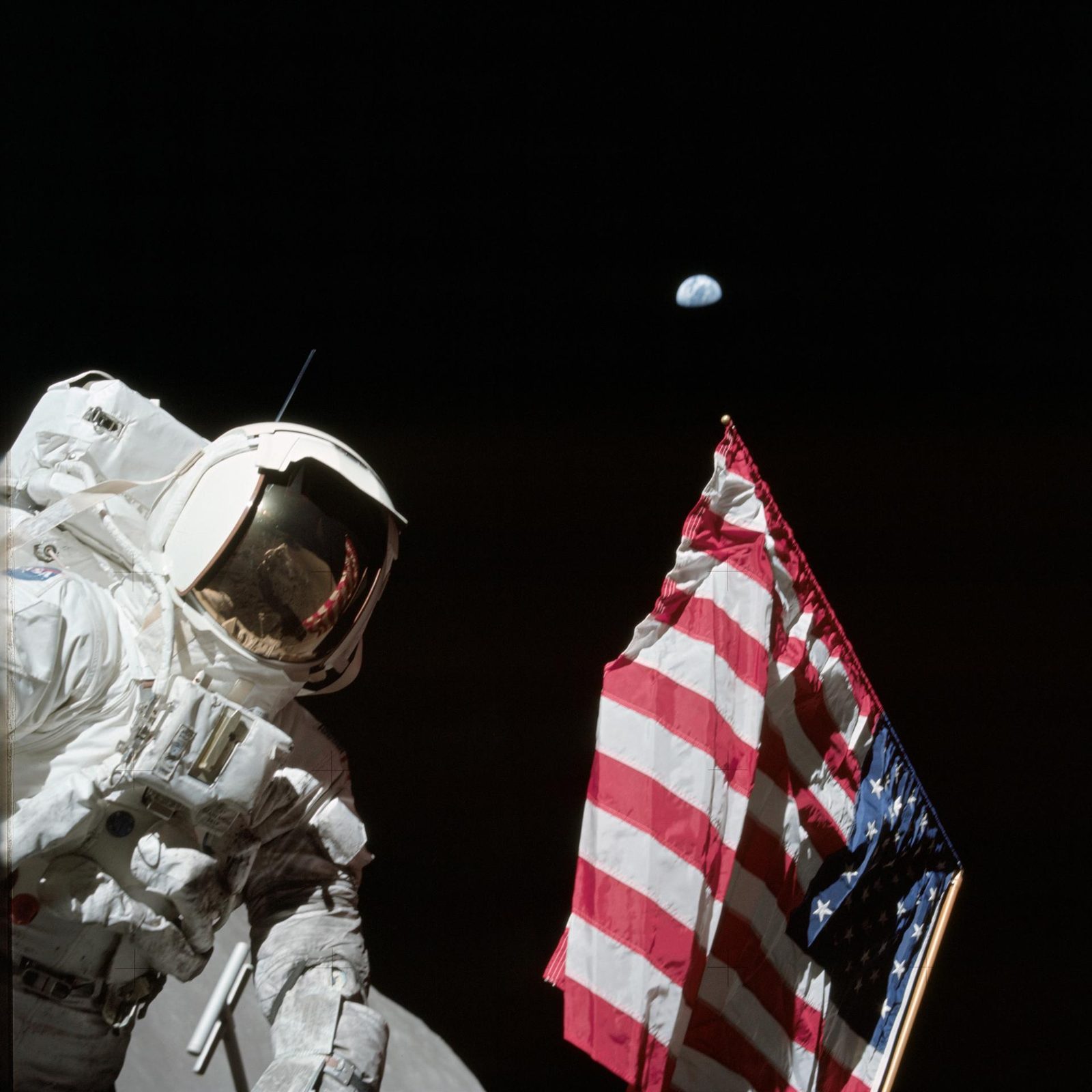 Un astronauta de pie junto a una bandera clavada en el suelo gris de la Luna, con una ladera clara detrás.