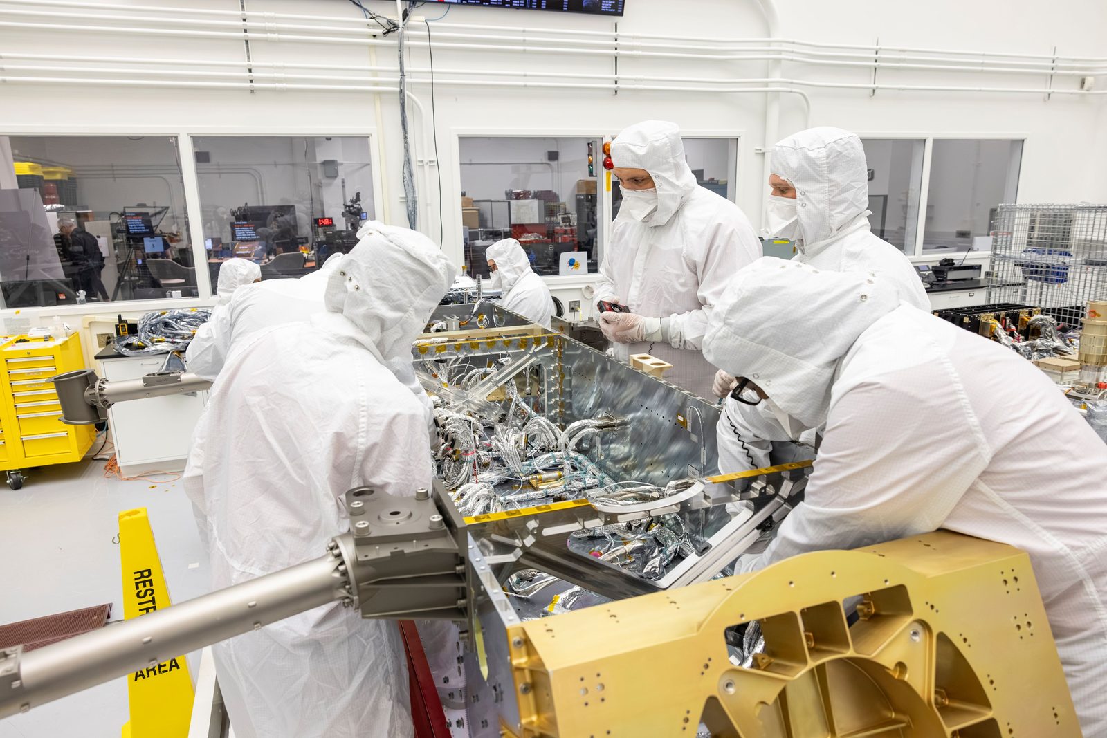 Varios técnicos con bata y cofia de sala limpia trabajan inclinados sobre el fuselaje abierto de un vehículo espacial, manipulando mazos de cables en su interior.