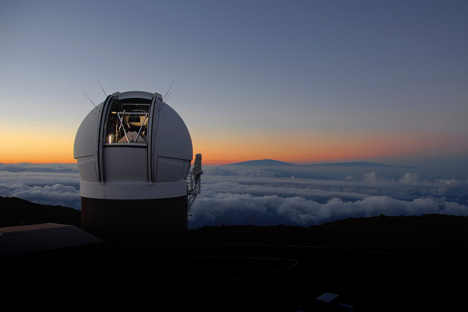 Una cúpula blanca de observatorio con el postigo abierto, sobre una cima, con el cielo al atardecer y un mar de nubes por debajo.