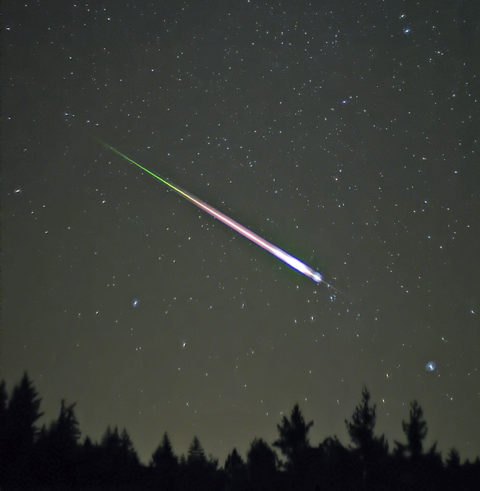 Un bólido brillante cruza en diagonal un cielo estrellado sobre la silueta de unos abetos, con una estela verde y una cabeza blanca y azulada.