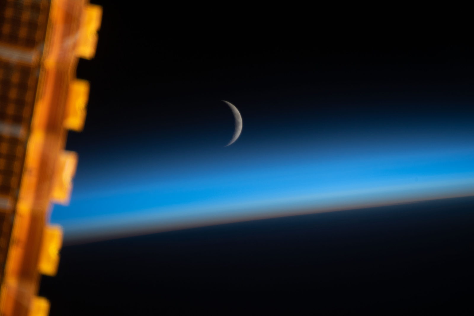 Una Luna creciente muy fina sobre el borde azul de la atmósfera terrestre, con un panel solar de la Estación en primer plano.