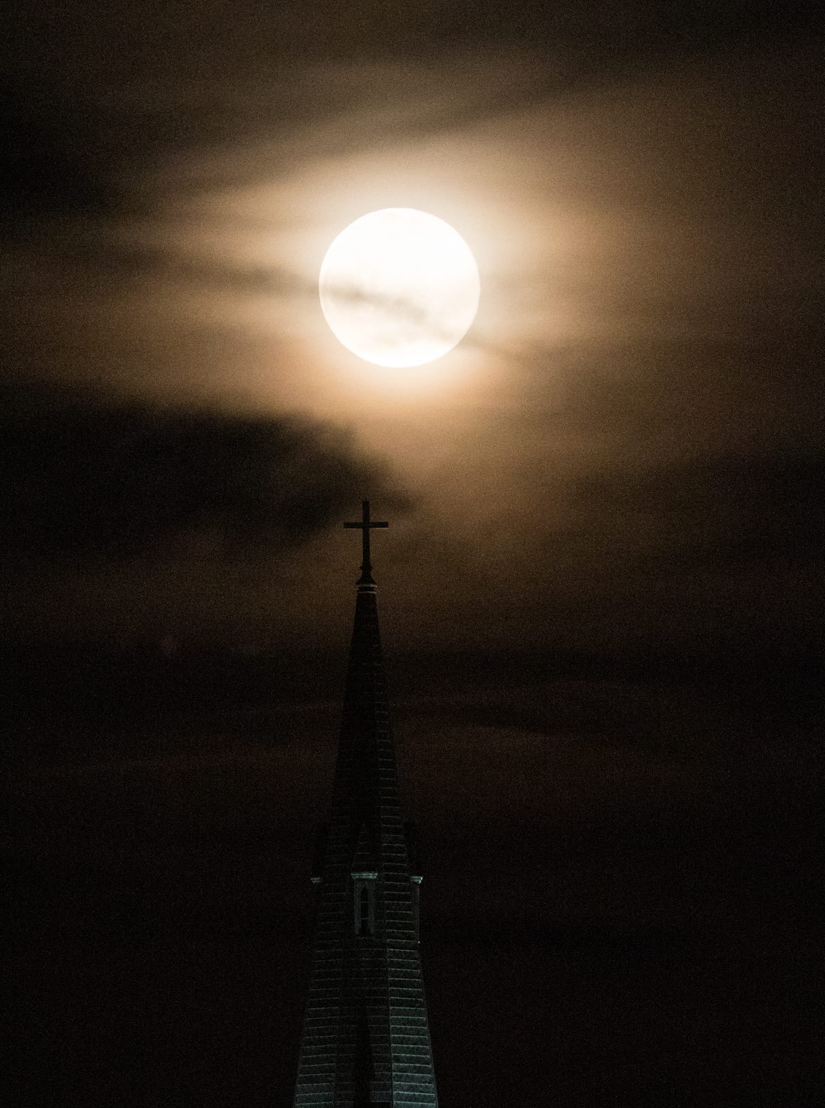 La Luna llena, grande y anaranjada, asomando justo detrás de la aguja de una iglesia recortada en negro.
