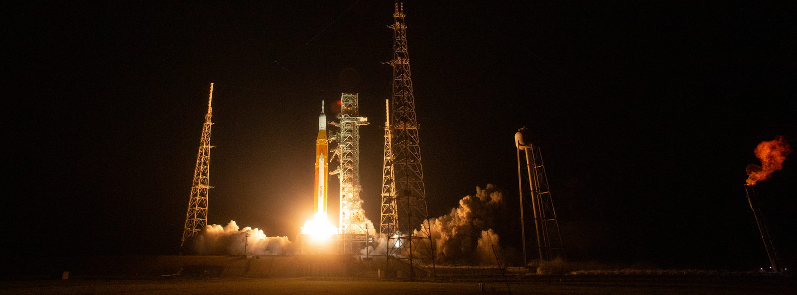 Un cohete despega de noche envuelto en una columna de fuego naranja que ilumina el vapor y el agua de alrededor.