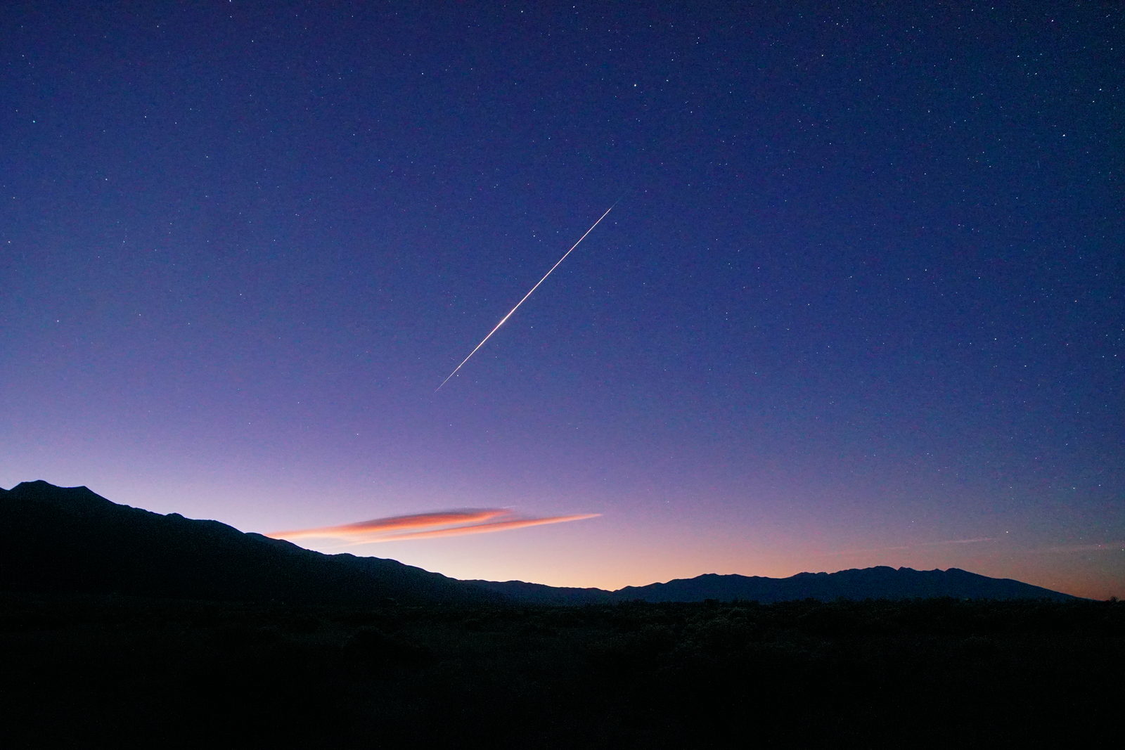 Una estela de meteoro sobre un cielo que ya clarea al amanecer, con nubes lenticulares recortadas sobre el horizonte.