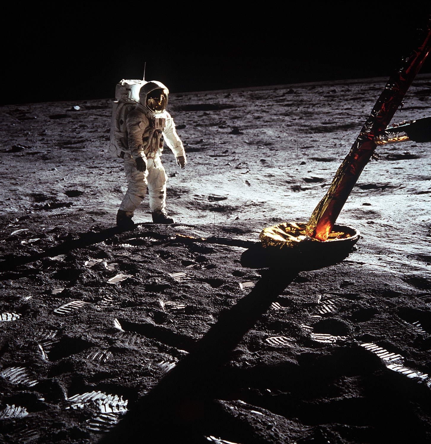 Un astronauta con traje blanco camina sobre el suelo gris de la Luna junto a una pata del módulo lunar; sus huellas quedan marcadas en el polvo.